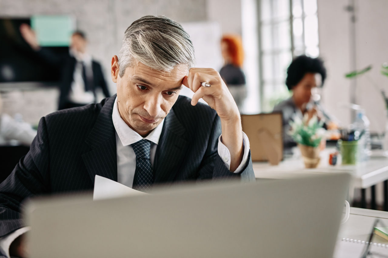 Pensive businessman thinking how to solve problems while doing paperwork in the office. There are people in the background. 