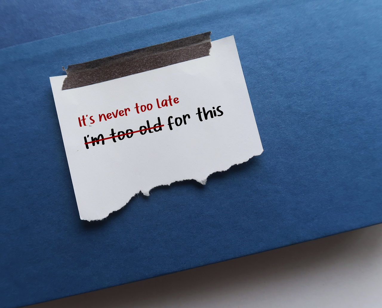 Stick torn note on blue text books with text written I AM TOO OLD FOR THIS, changed to IT IS NEVER TOO LATE FOR THIS - to replace negative self criticism to positive self talk with growth mindset